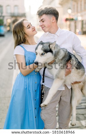 Young joyful couple in casual clothes, posing with adorable husky dog in the street of beautiful morning city, looking each other and smiling