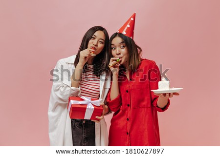 Tanned Asian women blow out party horns on isolated. Cute woman in red blouse and party hat holds birthday cake. Young lady in stylish outfit poses with gift box on pink background.