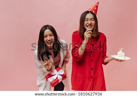 Joyful brunette Asian women laugh on pink isolated background. Lady in party hat and red blouse holds birthday cake. Girl in striped shirt poses with gift box.