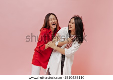 Happy young women in trendy oversized shirts laugh on isolated. Brunette Asian girls in stylish outfits smile and have fun on pink background.
