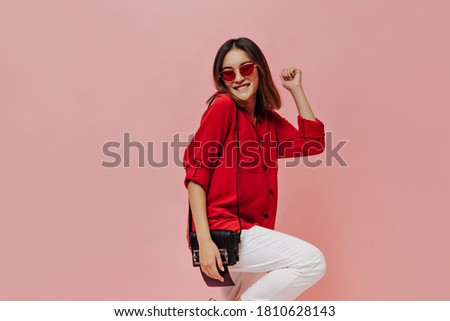 Cool young attractive Asian woman in red shirt, white pants and sunglasses smiles, holds handbag and dances on pink background.