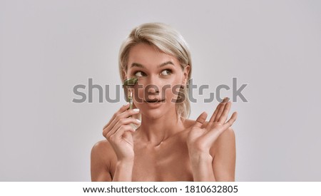 Portrait of young woman with perfect glowing skin making face while using jade facial roller for skin care and beauty treatment, posing isolated over grey background, Front view, Web Banner