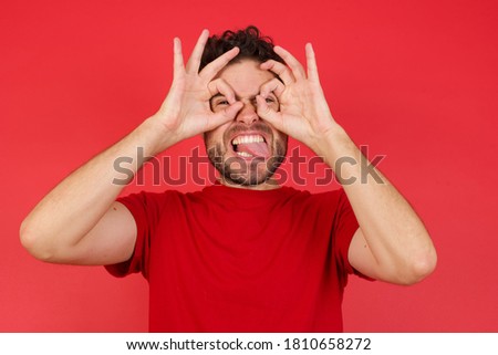 Young handsome caucasian man wearing t-shirt over isolated red background doing ok gesture like binoculars sticking tongue out, eyes looking through fingers. Crazy expression.