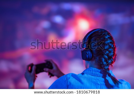 The girl gamer is actively playing games with her back, against the background of a 4k TV, or a larger screen. Gaming, streaming, joystick and passing the game
