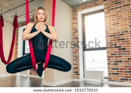 Caucasian blonde woman with closed eyes doing antigravity yoga meditating while hanging in lotus pose on red hammock in yoga fitness hall with panoramic windows