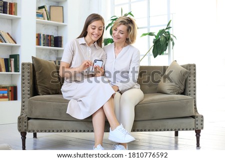 Middle aged mother and adult daughter hugging, using phone together, watching video or photos, sitting on cozy couch at home.