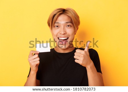 Close-up of happy and satisfied asian handsome guy, showing credit card and thumbs-up in approval, smiling amazed, standing yellow background