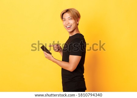Portrait of pleased smiling handsome asian man, standing in profile and pointing finger at smartphone, recommend app, standing yellow background