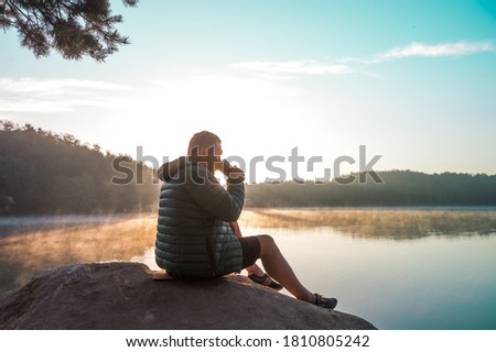Man enjoying a cup of coffee in sun rays outdoors in beautiful nature. Hiker in down jacket sitting on top of a mountain and enjoying sunrise.