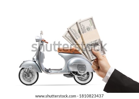 Hand of a young man in a suit holding stacks of us dollar money and a silver vintage scooter in the back isolated on white background