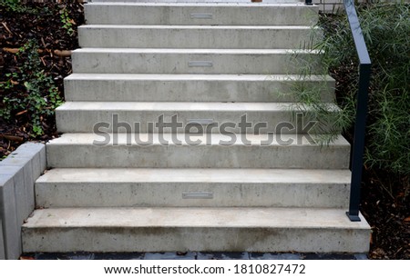 bright monolithic concrete park staircase with stair lighting with metal railing black interlocking paving, bushes slope