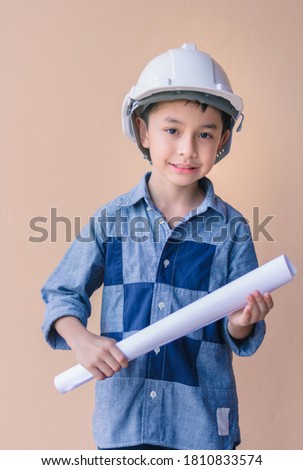 Asian boy in costume of architect hand touching helmet and holding blueprint isolate on orange background