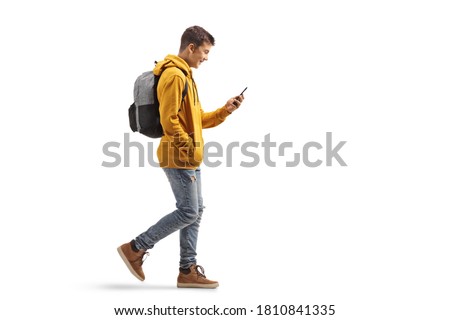 Full length profile shot of a male teenage student walking and looking at a mobile phone isolated on white background