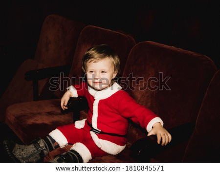 little boy child in a red costume of santa claus and with blond hair in felt boots on a dark black background. 