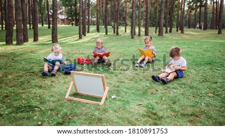New normal back to school. Kindergarten and elementary scholars sitting on grass at open air class during pandemic. Safe hybrid education, social distance rule, new schooling guidance