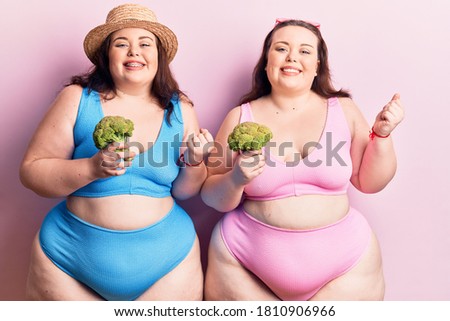 Young plus size twins wearing bikini holding broccoli screaming proud, celebrating victory and success very excited with raised arm 
