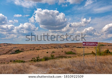 Brown Tourist information board (inscription in Cyrillic and Latin) in a field pointing to a rock church from the tenth century - a field yellowed by land with a blue sky and white fluffy clouds