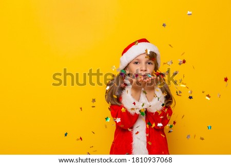 Portrait of a beautiful little girl wearing Santa Claus costume, having fun while blowing colorful star-shaped confetti on yellow colored background