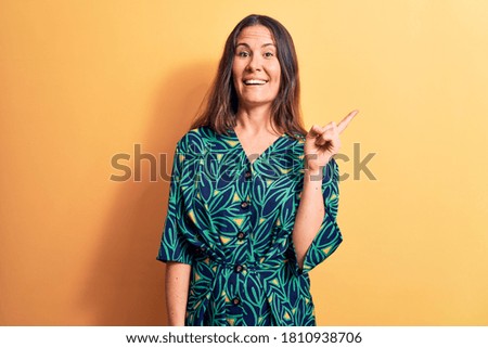 Young beautiful brunette woman wearing casual floral dress standing over yellow background with a big smile on face, pointing with hand and finger to the side looking at the camera.