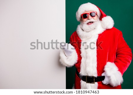 Photo of retired stylish grandpa grey hair beard direct finger white empty board promoting funny event wear red santa x-mas costume coat gloves sunglass cap isolated green color background