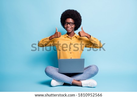 Full length photo dark skin high school girl sit floor legs crossed study remote laptop enjoy seminar watch show thumb up sign wear yellow shirt denim jeans isolated blue color background