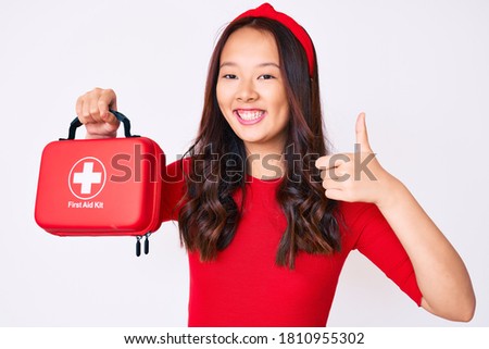 Young beautiful chinese girl holding first aid kit smiling happy and positive, thumb up doing excellent and approval sign 
