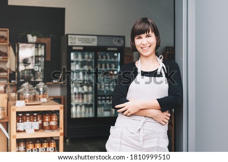 Portrait of owner of sustainable small local business. Shopkeeper of zero waste shop standing on interior background of shop. Smiling young woman in apron welcoming at entrance of plastic free store Royalty-Free Stock Photo #1810999510