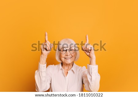 Portrait of smiling old woman with short gray hair and round glasses wearing white blouse, pointing fingers up. Senior woman isolated over orange background Royalty-Free Stock Photo #1811010322