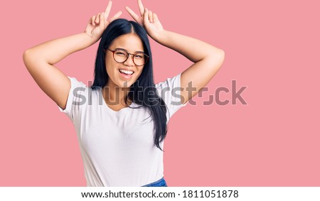 Young beautiful asian girl wearing casual clothes and glasses posing funny and crazy with fingers on head as bunny ears, smiling cheerful 