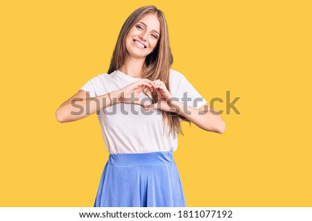 Young beautiful blonde woman wearing summer style smiling in love doing heart symbol shape with hands. romantic concept. 