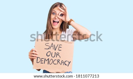 Young beautiful blonde woman holding save our democracy protest banner smiling happy doing ok sign with hand on eye looking through fingers 