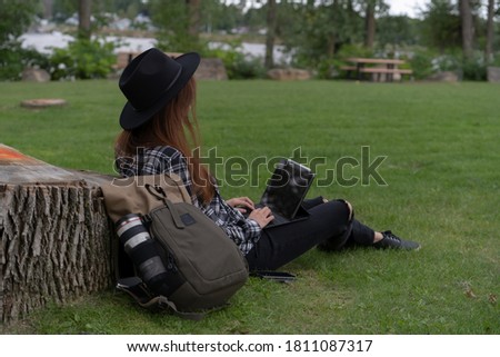 woman covering her face dressed in a plaid shirt and black hat sitting in the middle of a park next to her photography suitcase working with her laptop on her lap