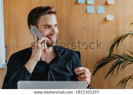 Business and communication concept. Handsome young man sitting at office desk with laptop computer and talking on mobile phone.
