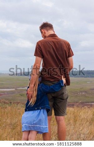 Father and daughter standing on a hill with nature lanscape back view. Little blonde girl with hugging with dad outdoor. Loving child embrace her daddy. Father's Day family greeting card faceless.
