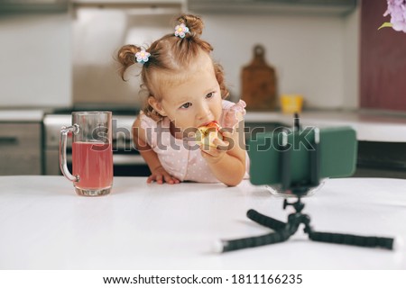 Portrait of a Cute little girl sitting at the kitchen table, eating an apple and watching a video on her phone, space for text