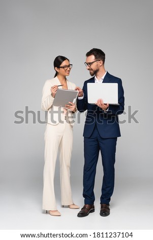 full length view of businessman with laptop pointing with finger near asian colleague holding folder on grey Royalty-Free Stock Photo #1811237104