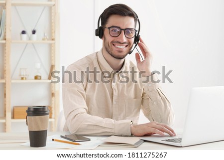 Young man from customer service wearing beige shirt, eyeglasses and headset supporting client via voice call, smiling happily at camera