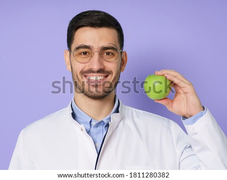 Close-up portrait of smiling nutritionist or dentist doctor wearing white coat, holding fresh green apple with hand, isolated on purple background