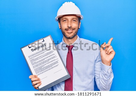 Young handsome man wearing architect hardhat holding contract document smiling happy pointing with hand and finger to the side 
