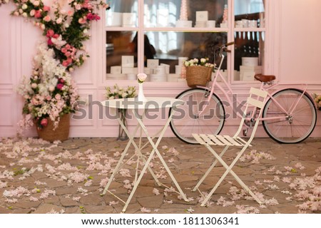 Empty cafe terrace with white table and chair. Pink exterior of the cafe restaurant. interior Street cafe. Cozy street with flowers and French-style cafe table. Decor facade of coffeehouse with bike.  Royalty-Free Stock Photo #1811306341