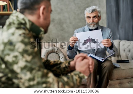 Mature psychologist holding picture with ink stain, Rorschach Inkblot in front of military man during therapy. Soldier suffering from depression, psychological trauma. PTSD concept. Focus on picture
