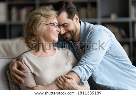 Smiling mature woman and adult son cuddling, enjoying tender moment, sitting on cozy couch in living room, happy beautiful elderly mother wearing glasses and young man hugging, two generations Royalty-Free Stock Photo #1811335189