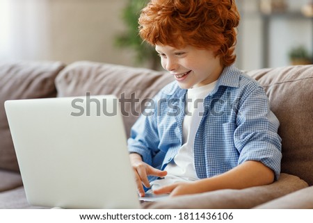 Full body cheerful redhead boy in casual clothes  smiling and playing game on laptop while sitting on soft couch at home
