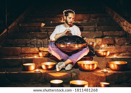 silhouette of a man playing handpan surrounded by Tibetan singing bowls and lighted candles sitting on the steps of the night city. Noise effect on the film