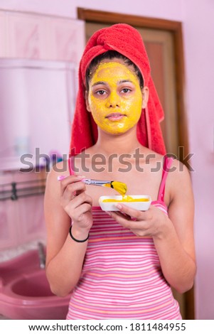Skin care - Beautiful girl applying Gram flour turmeric yellow face mask on face through brush. She is wearing red towel on head and looking at camera.