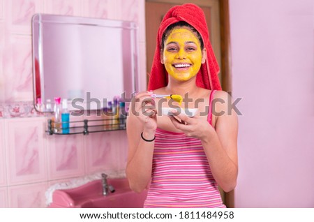 Skin care - Beautiful girl applying Gram flour turmeric yellow face mask on face through brush. She is wearing red towel on head and looking at camera with toothy smile.  Royalty-Free Stock Photo #1811484958