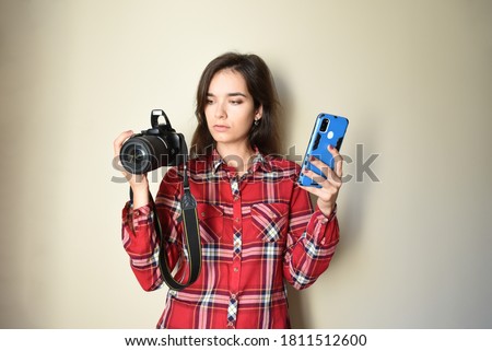Young brunette woman photographer in red shirt choosing between camera and mobile phone on beige background. Concept of choice and difference between mobile photography and camera. Copy space