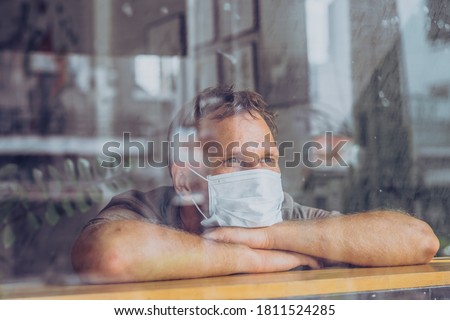 Man wear white protective mask, looking outside through window, afraid stress hope in eyes, recovery from the illness in home, self isolation due to global COVID 19 Coronavirus pandemic Royalty-Free Stock Photo #1811524285