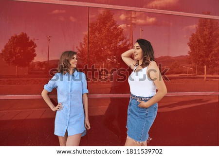 Two women posing on a red background. They're outdoors in a city.