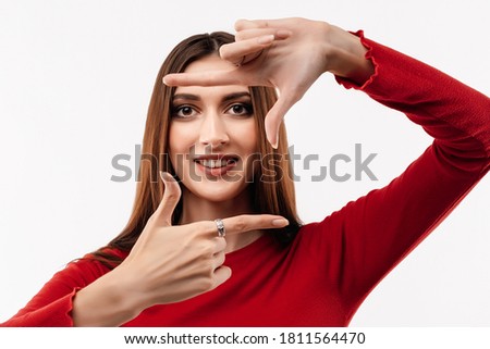 Photo of funny woman with long chestnut hair, wearing in casual red sweater, smiling and making photo frame with fingers. Human emotions, facial expression concept. Studio shot, white background